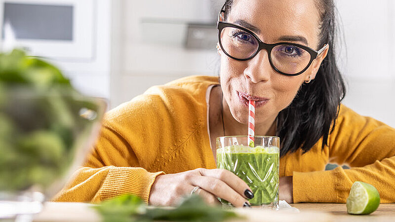 Frau blickt in die Kamera, während sie an einem grünen Shake nippt.