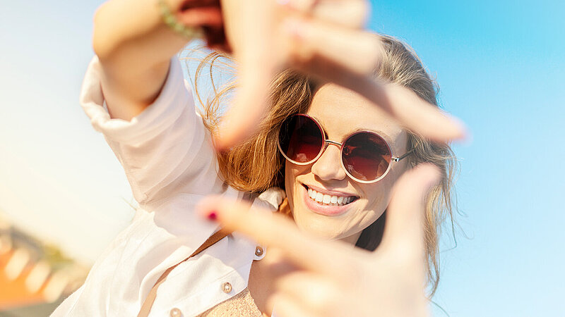 Frau mit Sonnenbrille blickt durch ein Quadrat, das sie mit ihrem Fingern formt. Im Hintergrund blauer Himmel