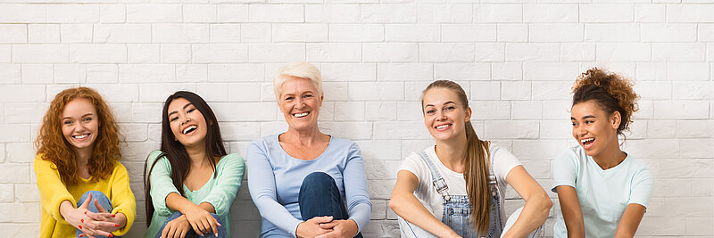 Fünf Frauen verschiedenen Alters sitzen vor einer Wand und wirken gut gelaunt