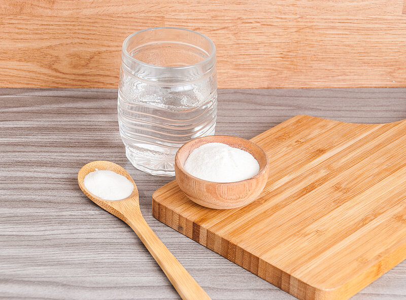 Auf einem Tisch liegt ein Holzbrett. Darauf eine Holzschale mit Kollagenpulver. Im Hintergrund ein Glas Wasser und ein Holzlöffel