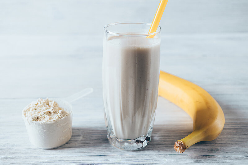 Ein Messlöffel Proteinpulver steht neben einem Glas Milch und einer Banane