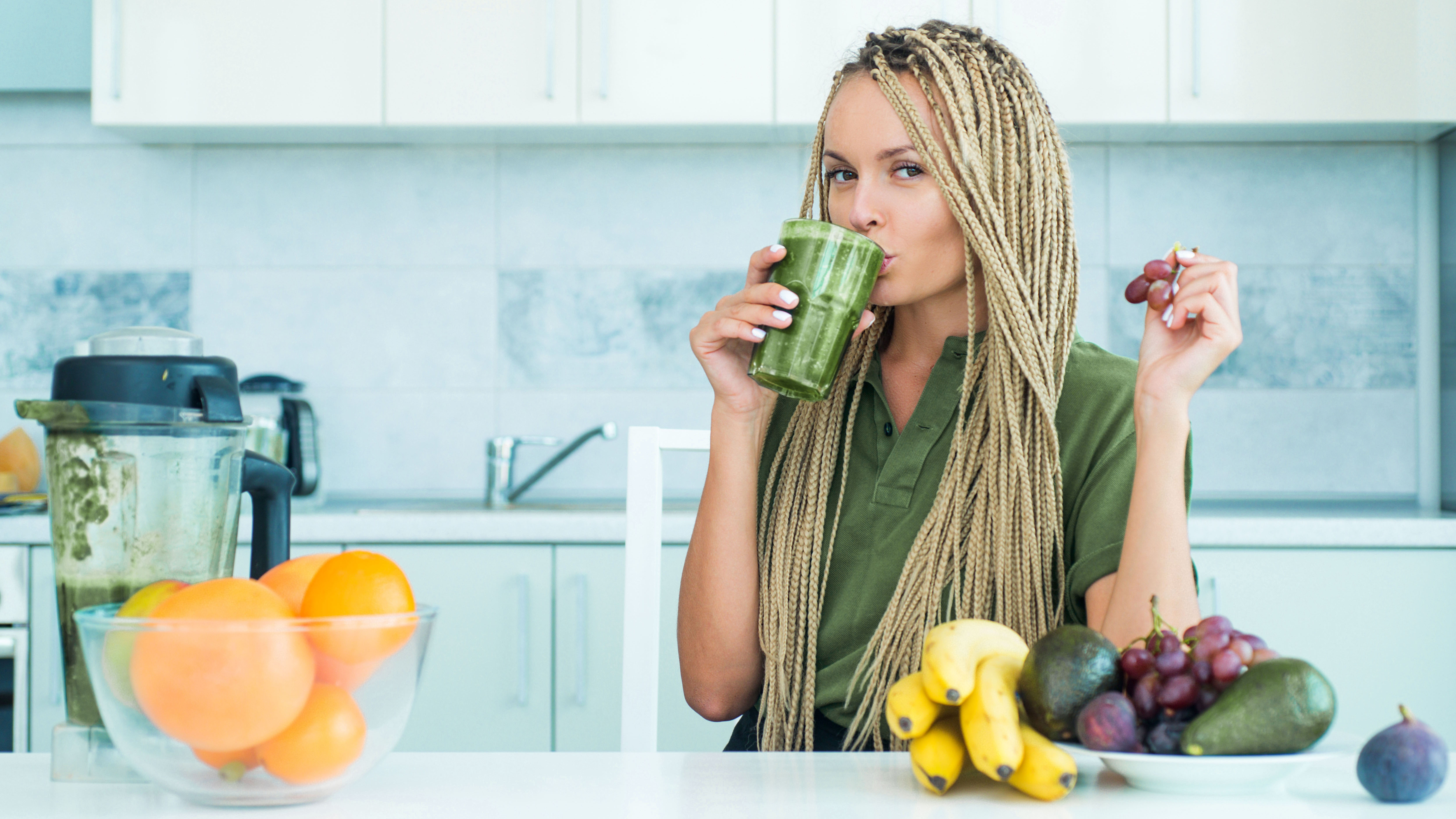 Junge Frau mit langen blonden Braids steht in der Küche und trinkt einen grünen Smoothie