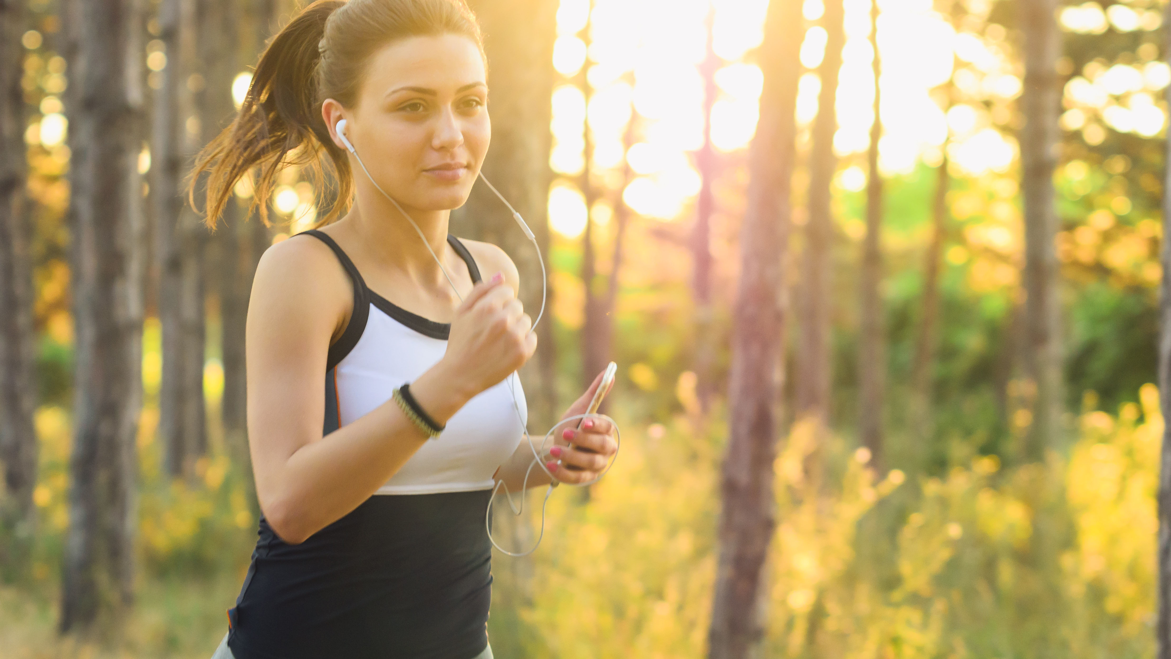 Junge Frau in schwarz-weißer Kleidung joggt von links nach rechts durchs Bild. Im Hintergrund ein Wald durch den das Sonnenlicht bricht