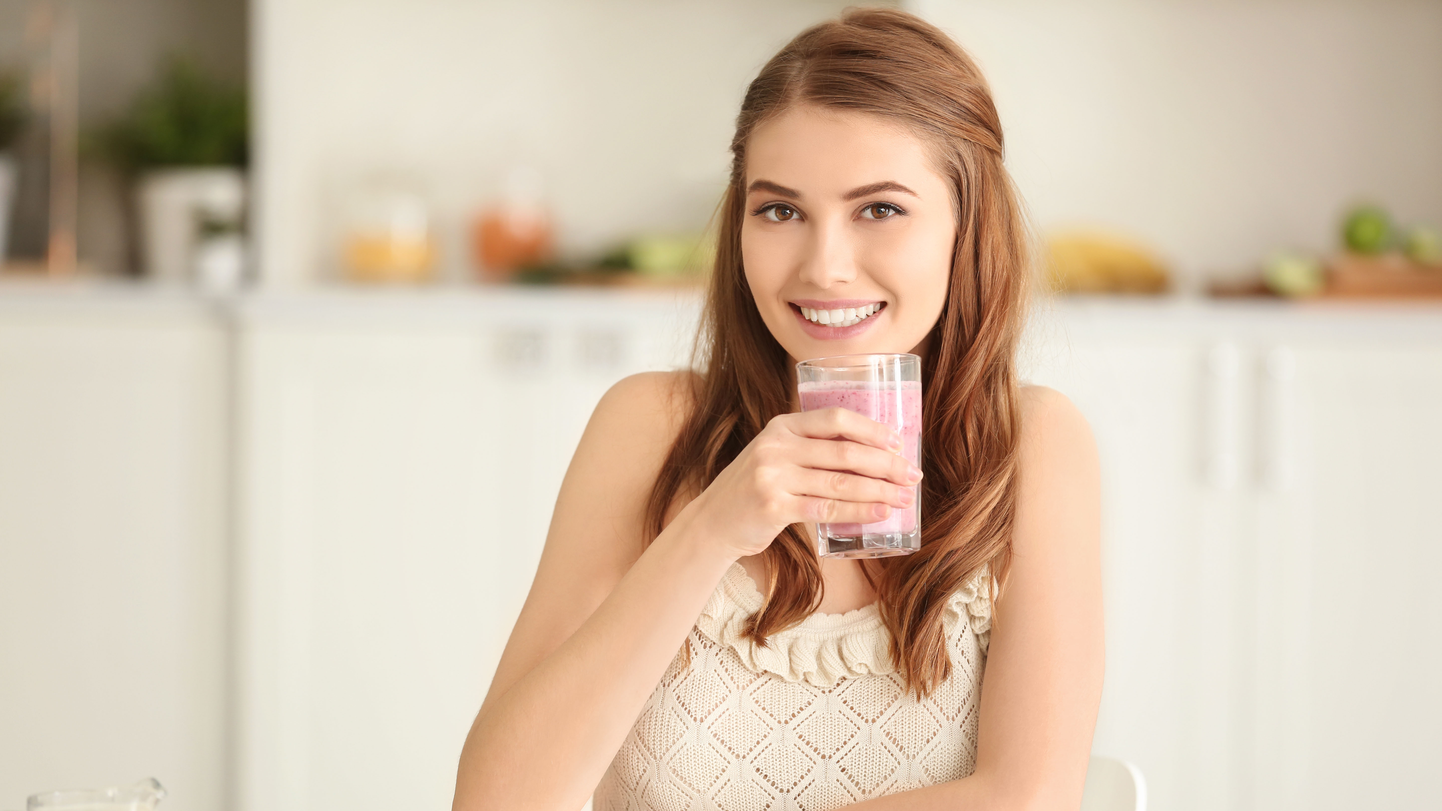 Junge rothaarige Frau Blickt in die Kamera und setzt zum trinken eines Frühstücksshakes an. Im Hintergrund ein verschwommenes Wohnzimmer in beige