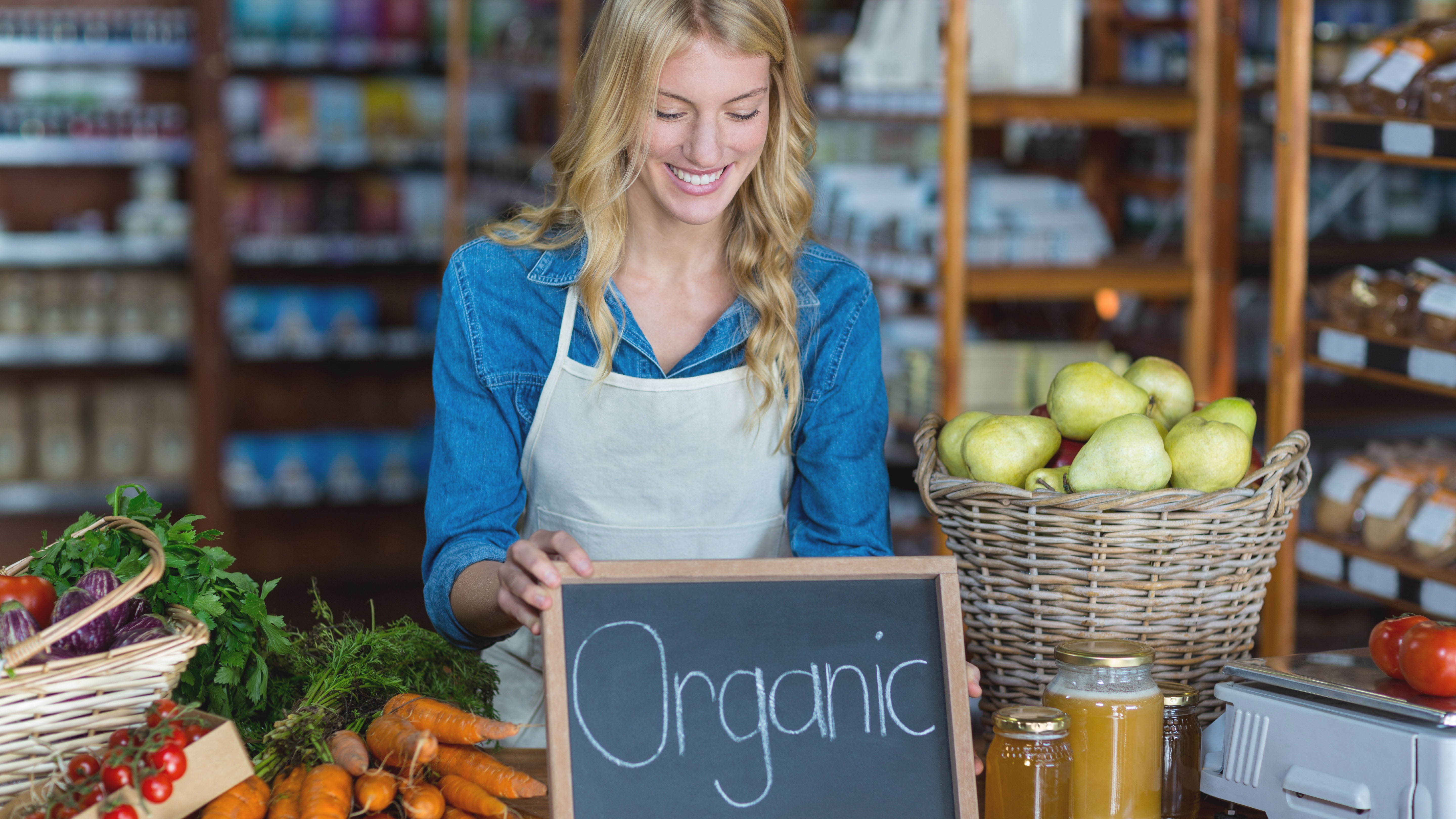 Blonde Frau in einem kleinen Hofladen trägt eine Schürze und Stellt eine Tafel auf, die mit Organic beschriftet ist