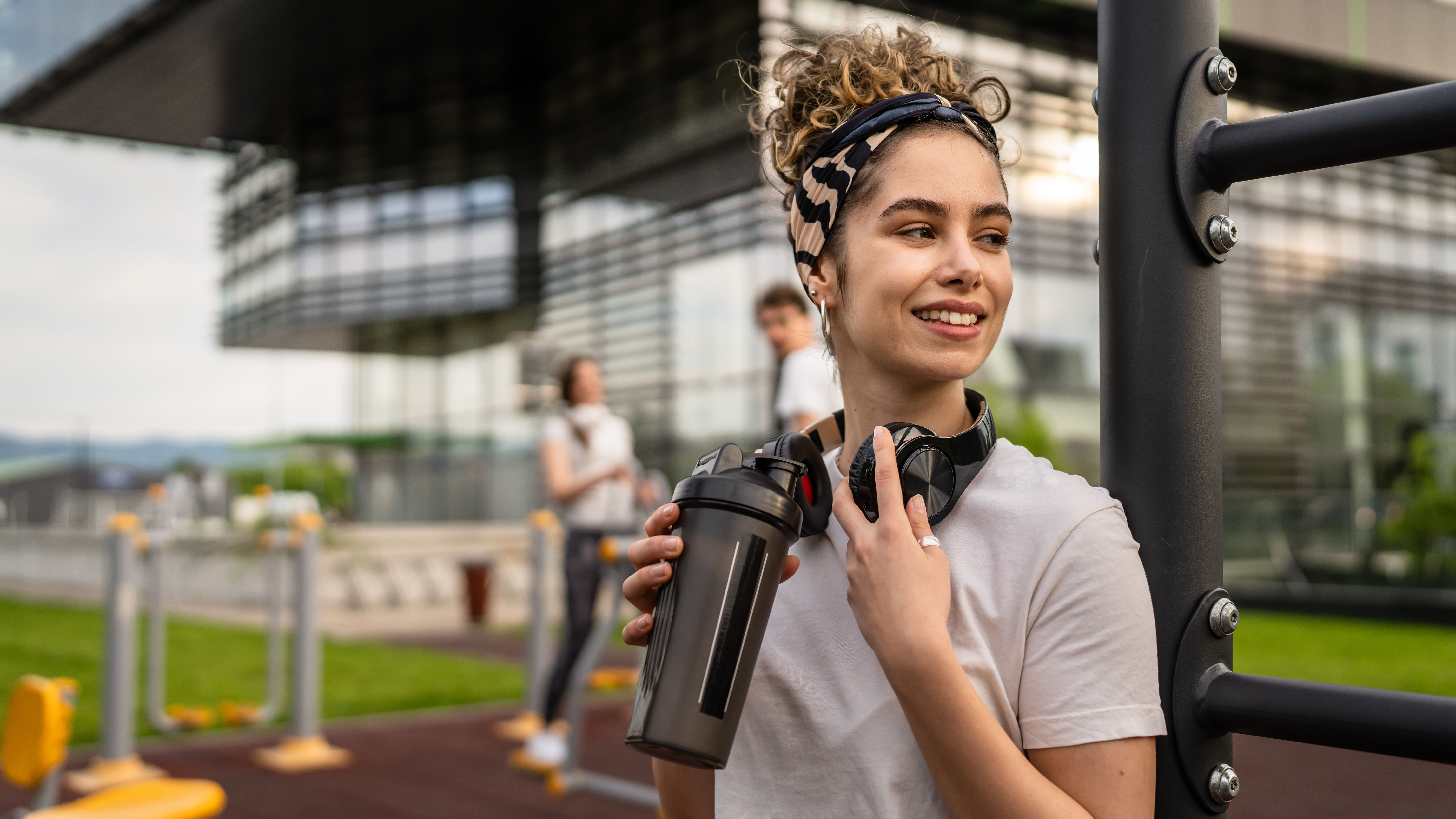 Junge Frau mit Haarband trinkt Clear Whey aus einem Shaker, im Hintergrund andere Sportler