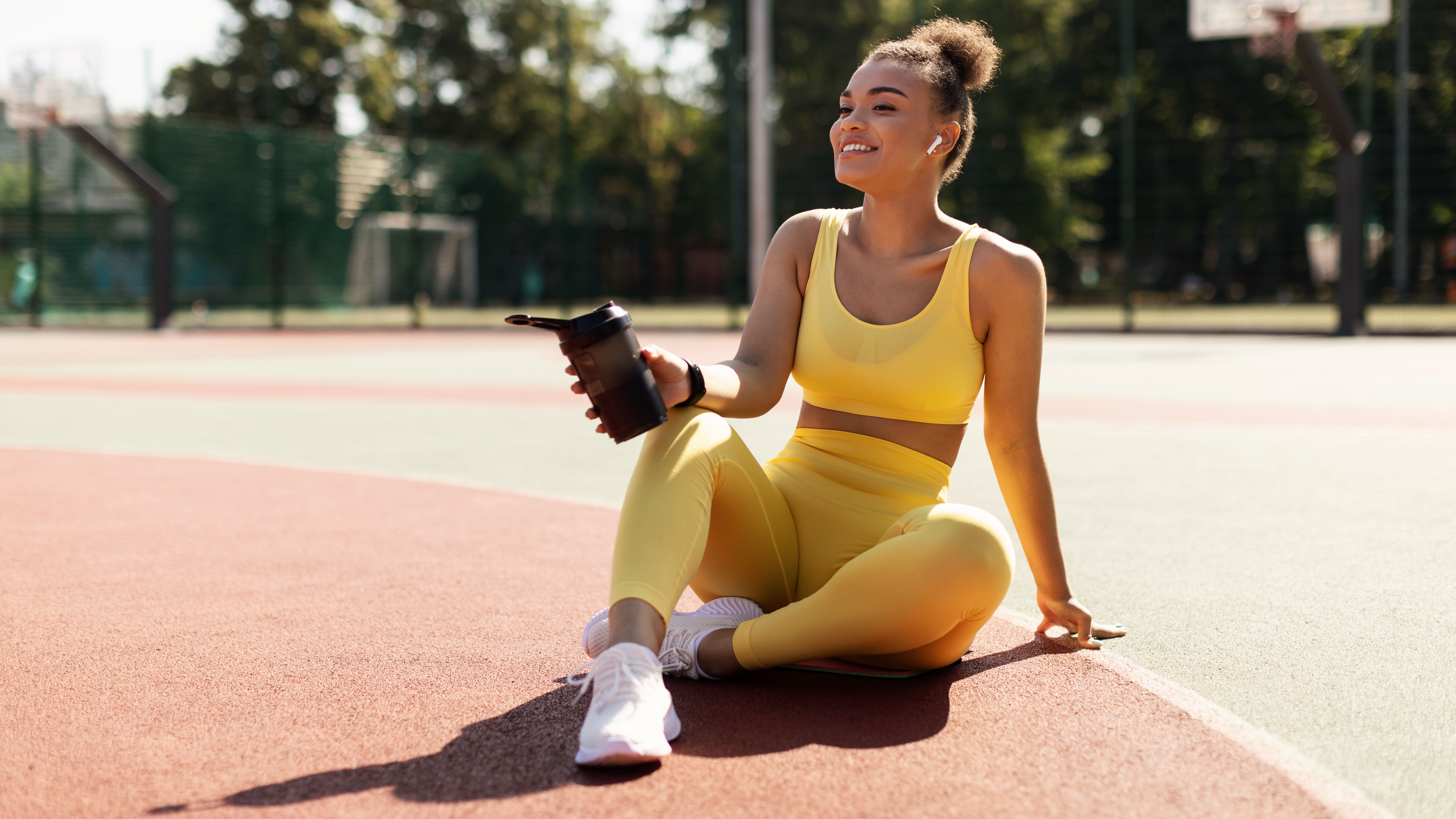 Junge Frau in gelben Sportklamotten sitzt mit Pre Workout Booster Shake auf dem Hartplatz