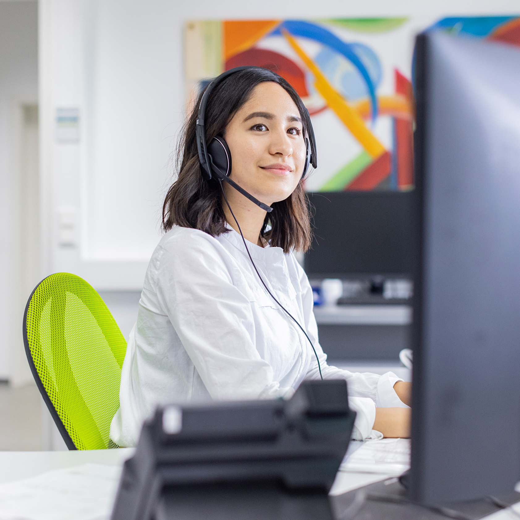 Junge Frau mit Headset sitzt auf einem grünen Stuhl im Büro und arbeitet am PC.