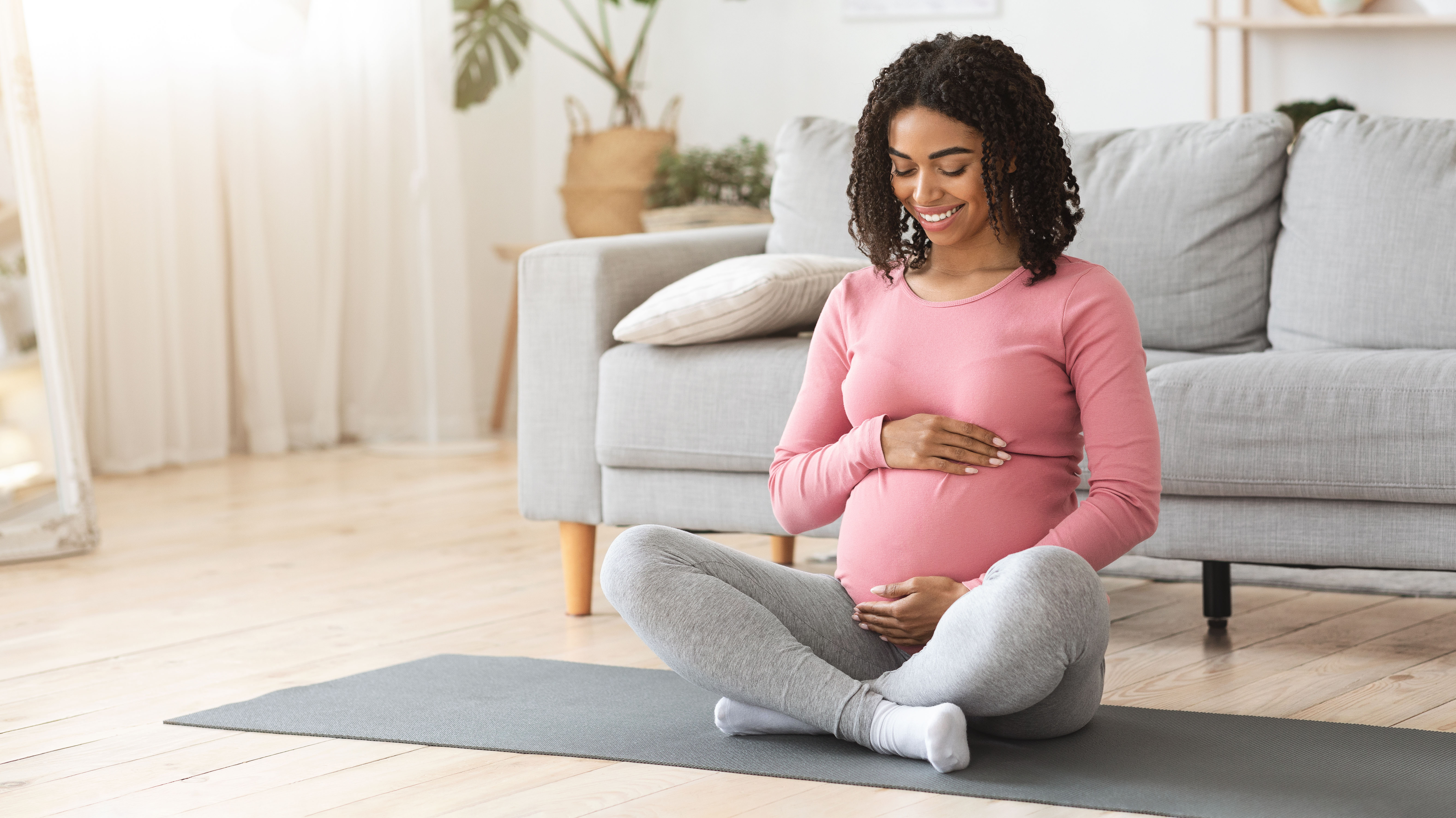 Schwangere Frau sitzt im Schneidersitz vor einer grauen Couch und blickt auf dem mit den Händen umfassten Bauch. 