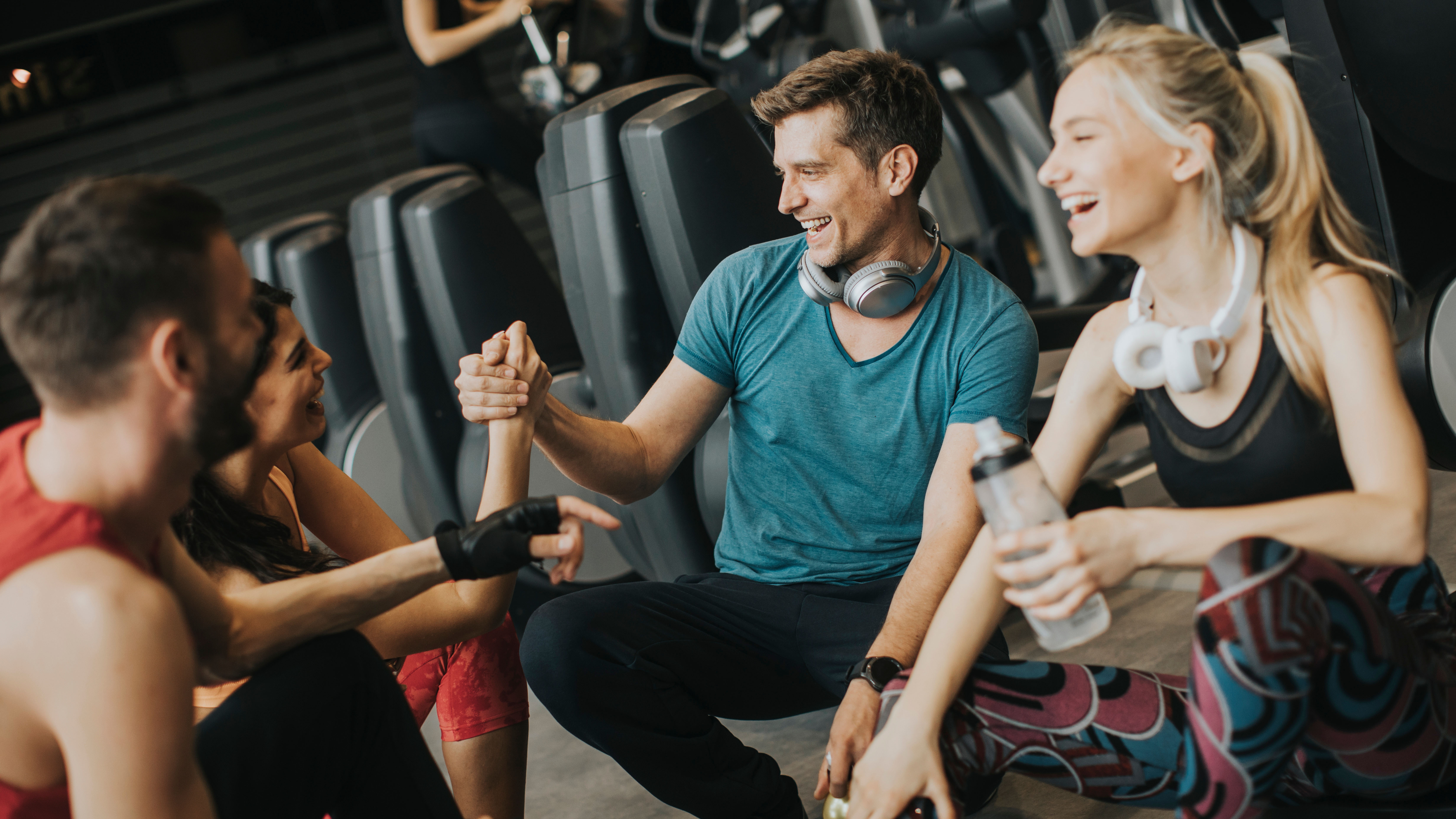 Gruppe von vier jungen Menschen sitzen nach dem Training gut gelaunt auf dem Boden des Fitnessstudios 