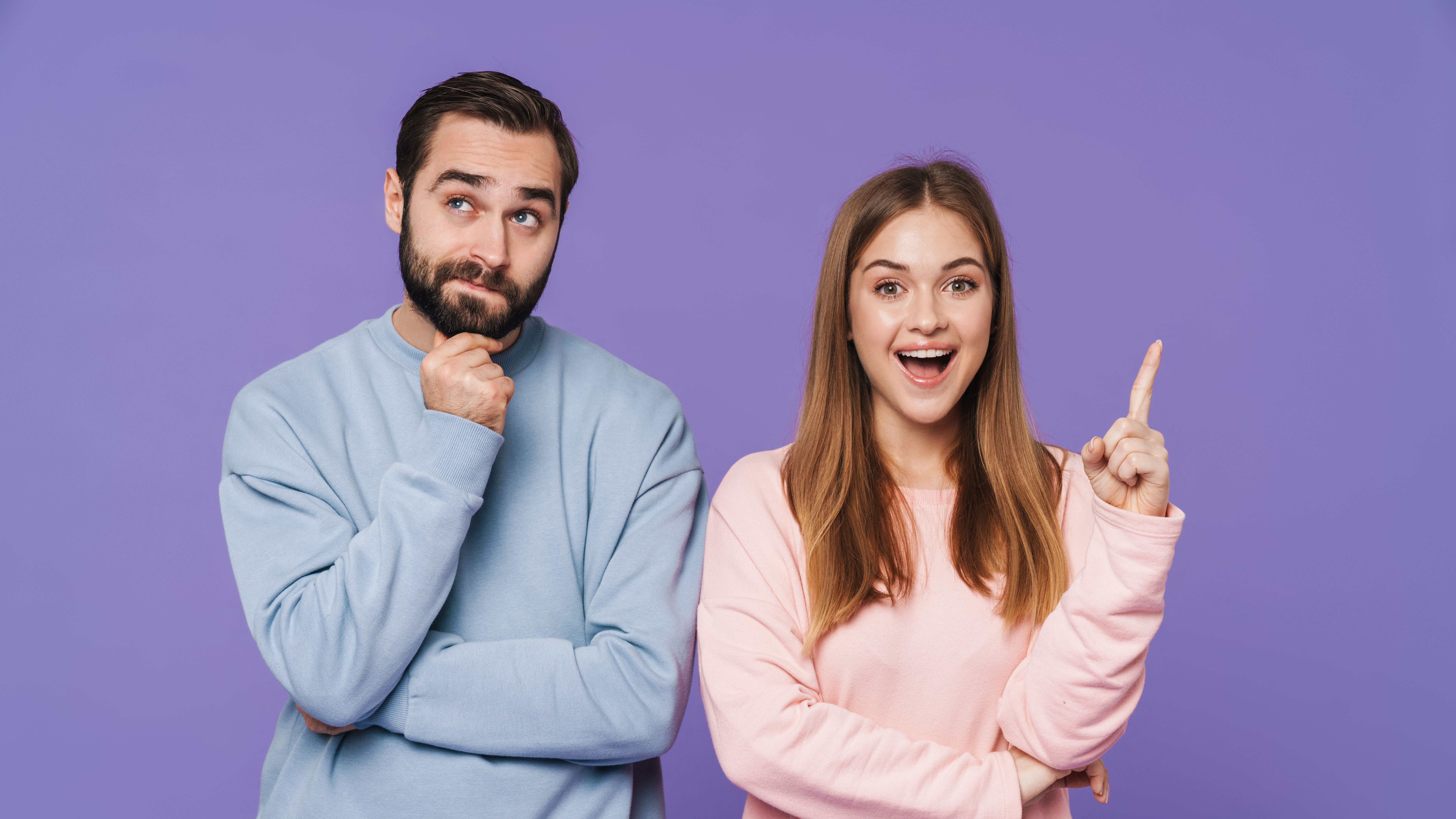 Mann in blauem Pulli und Frau im Rosa Pulli stehen vor lila Hintergrund. Er blickt Fragend in die Kamara, Sie hebt den Zeigefinger und schaut wissend. 