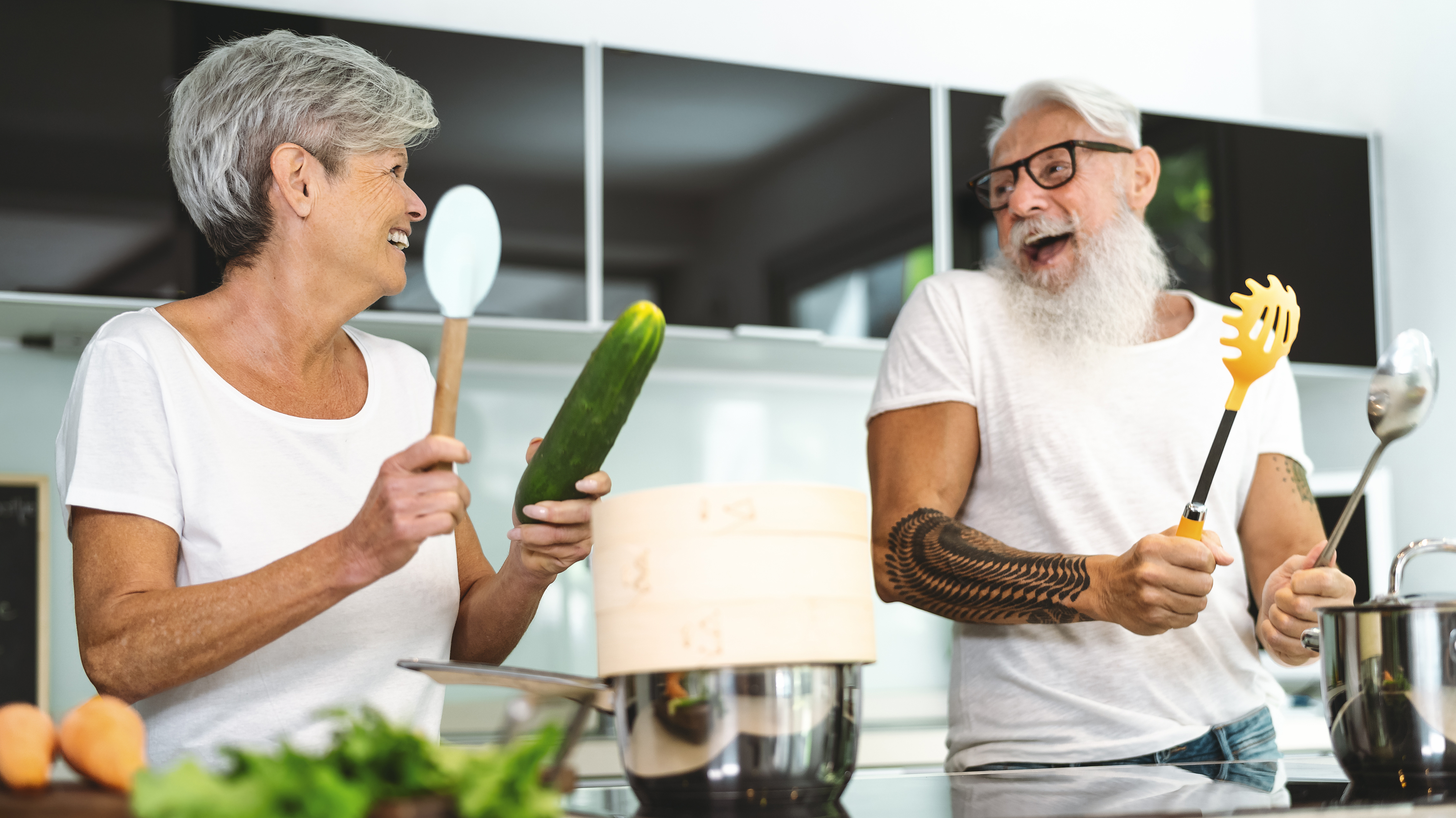 Älteres Paar hat viel Spaß beim gemeinsamen kochen und tanzen. Beide sind in weiß gekleidet, die Küche ist dunkel.