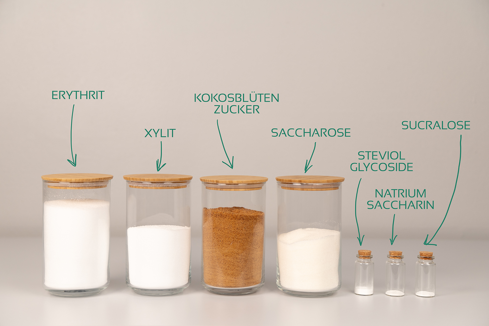 Vor einer grauen Wand stehen 4 große und 3 kleine Gläßer mit verschiedenen Süßungsmitteln nebeneinander. Die Füllmenge verdeutlicht die Süßkraft