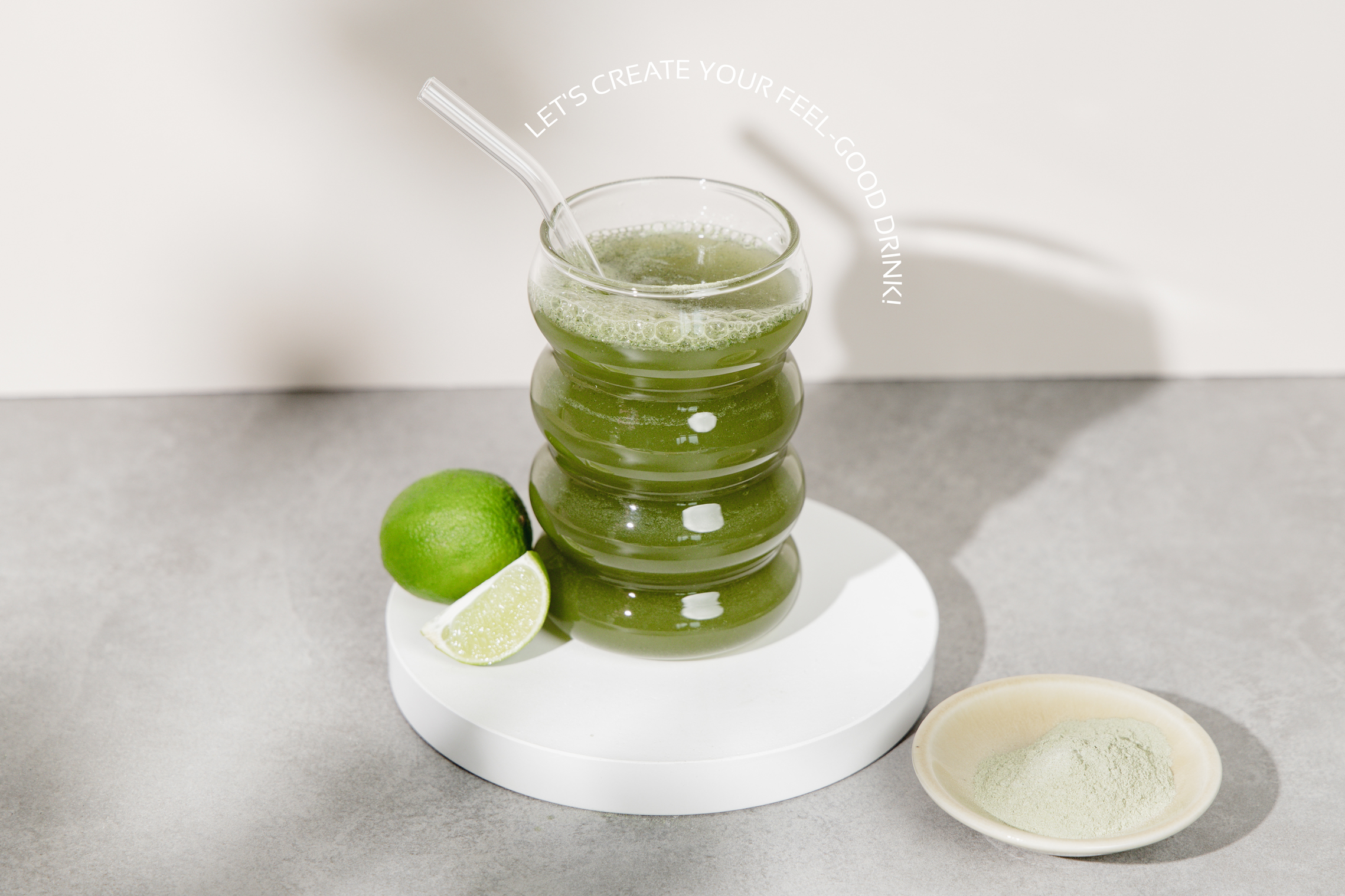 Ein Grüner Shake mit Limette steht vor grauem Hintergrund. Um das Glas steht "Let's create your Feel-Good-Drink"