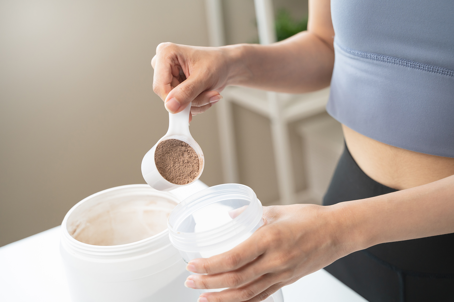 Person nimmt mit einem Messlöffel Proteinpulver aus einer Dose und gibt dieses in einen Shaker