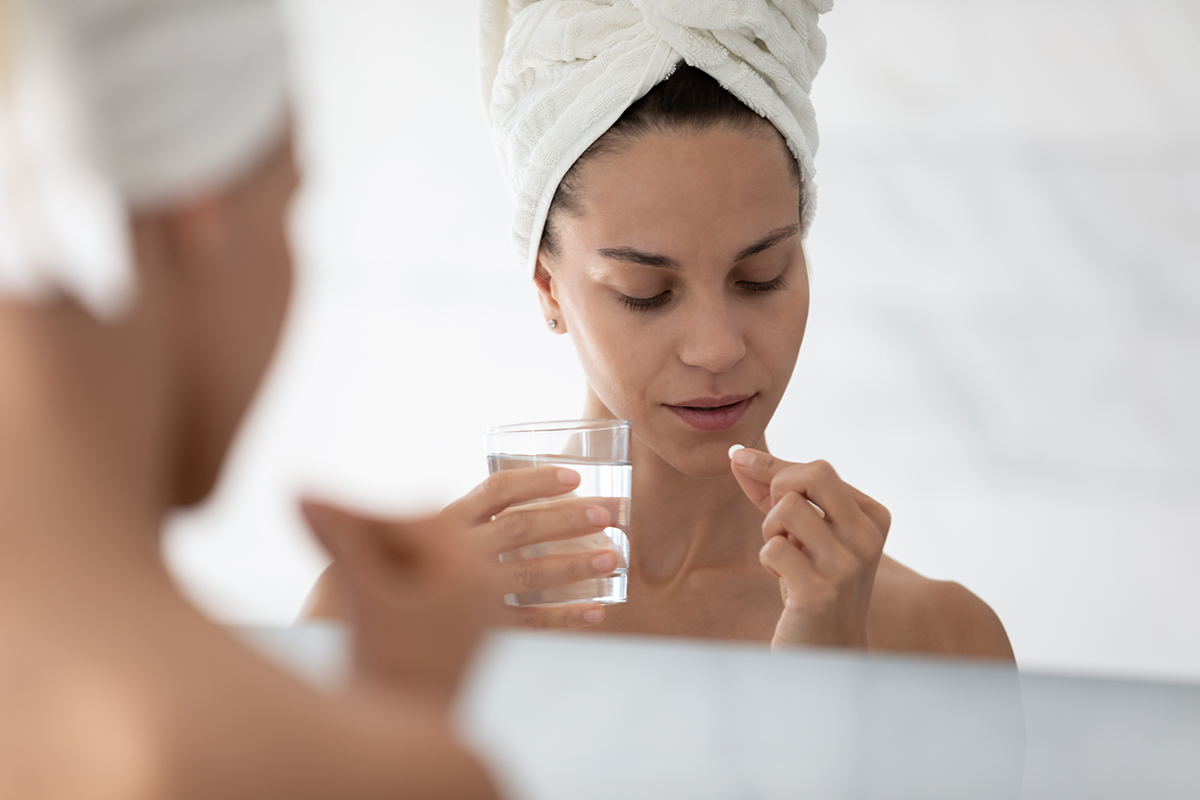 Frau mit Handtuchturban im Spiegel fotografiert während Sie ein Wasserglas in der linken und eine Kapsel in der rechten Hand hält um diese einzunehmen