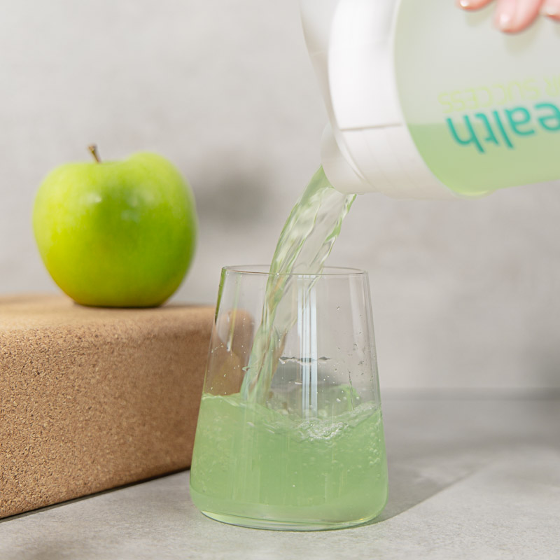 Private Label Energy Drink wird aus einem Shaker in ein Glas gegossen. Im Hintergrund Ein grüner Apfel auf einem Kork-Block