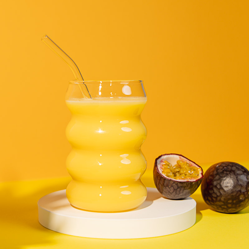 Gelber Hintergrund, davor ein gelber Drink im Glas mit Strohalm, daneben Maracuja