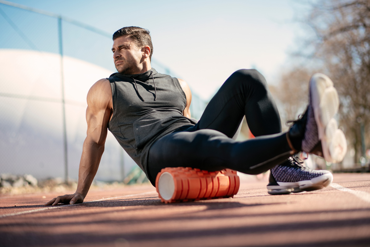 Sportlicher Mann sitzt auf einem Sportplatz mit einem Bein auf einer Faszienrolle um die Muskeln bei der Regeneration zu unterstützen