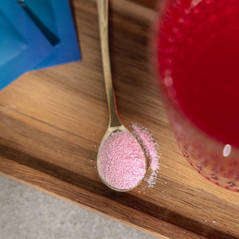 Isotonische Getränkepulver auf einem Löffel. Im Anschnitt des Bildes ein angemischter isotonischer Drink und ein Sachet