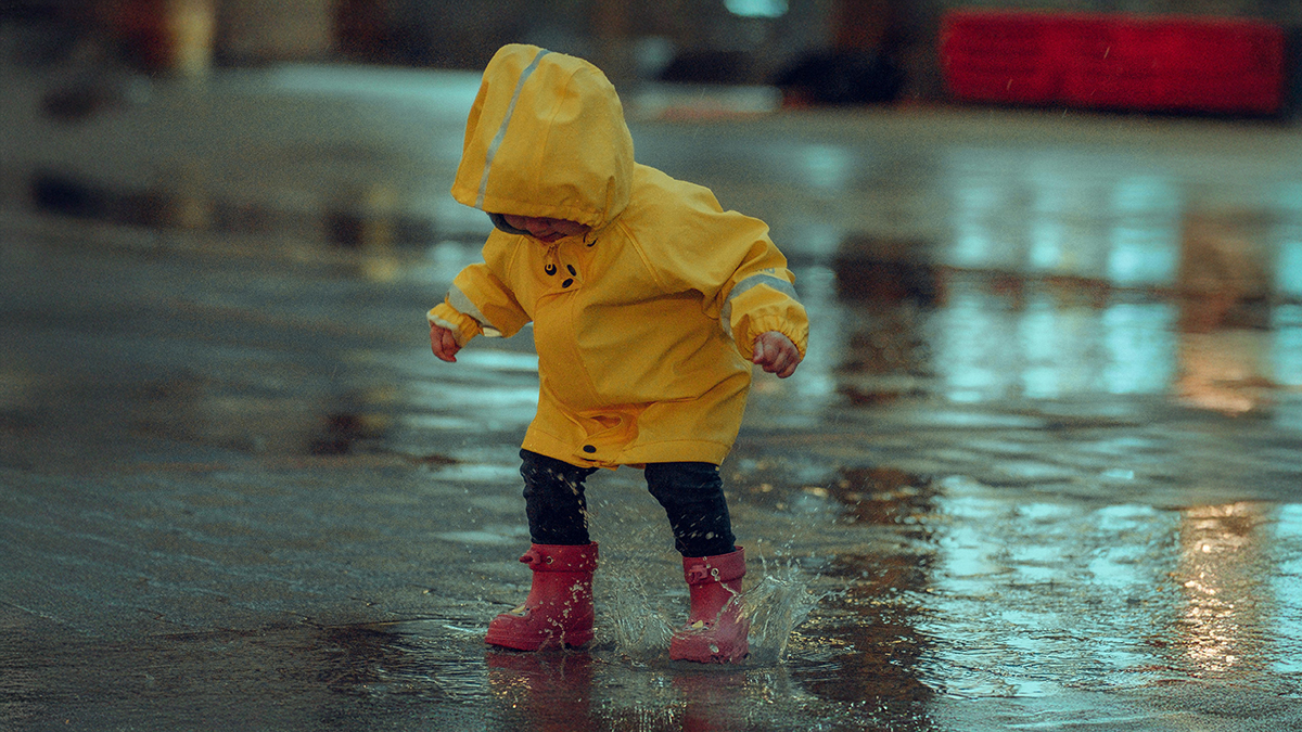 Kleines Kind mit Gummistiefeln und Regenjacke in einer Pfütze stehend