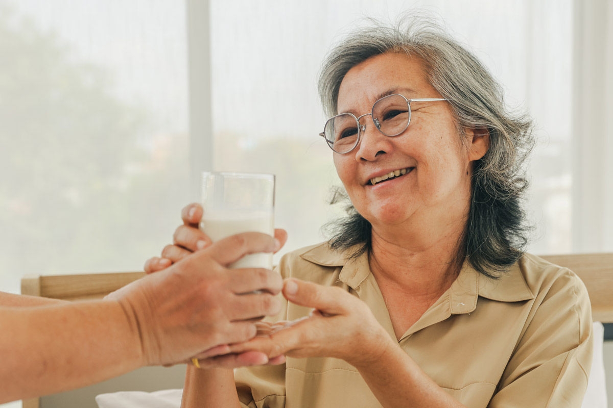 Ältere Dame nimmt ein Glas mit weißem Drink entgegen, das ihr von einer Hand aus dem linken Bildrand gereicht wird.