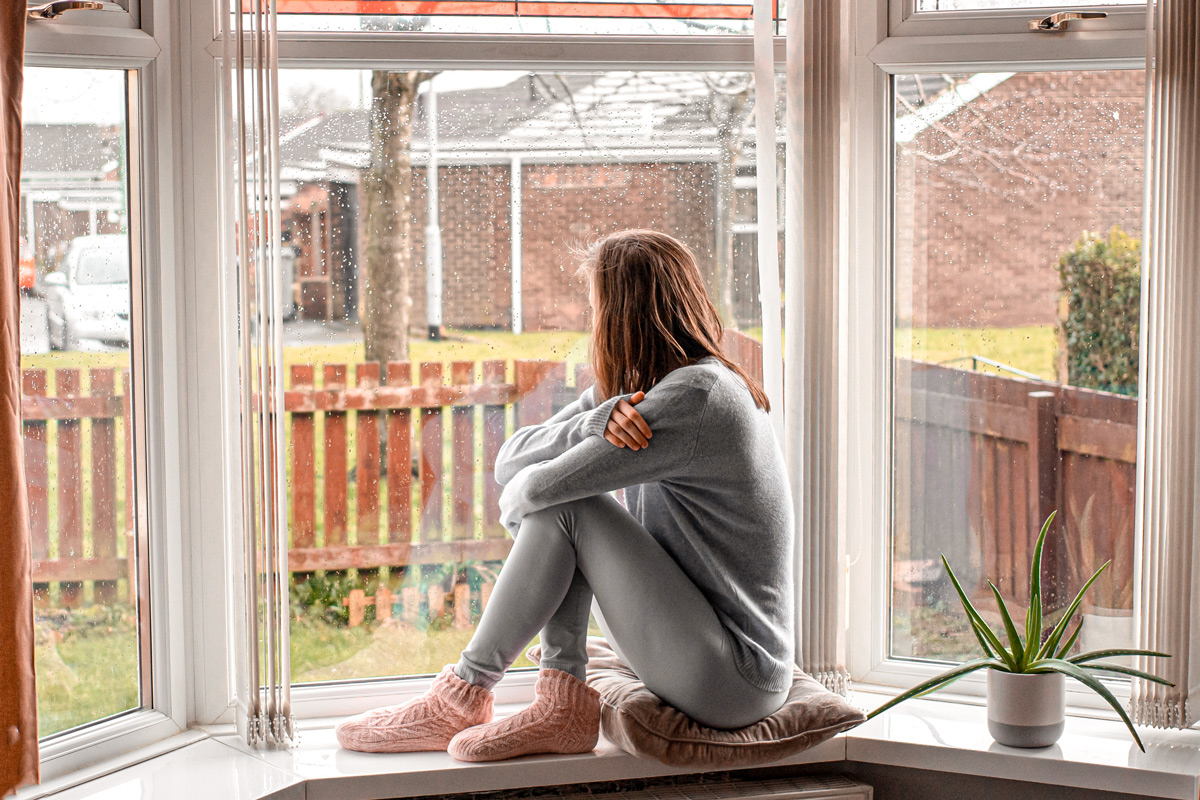 Frau sitzt auf Fensterbrett, auf dem leichte Regentropfen zu sehen sind, und blickt aus dem Fenster