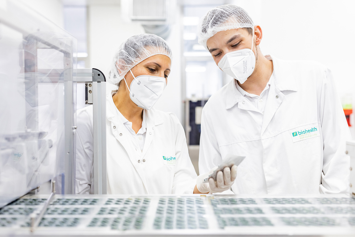 Mitarbeiterin aus der Produktion erklärt einem Quereinsteiger seine0n neuen Job in Münchberg beim Nahrungsergänzungsmittel Hersteller. Beide tragen weiße Produktionskleidung mit Masken und Haarnetzen